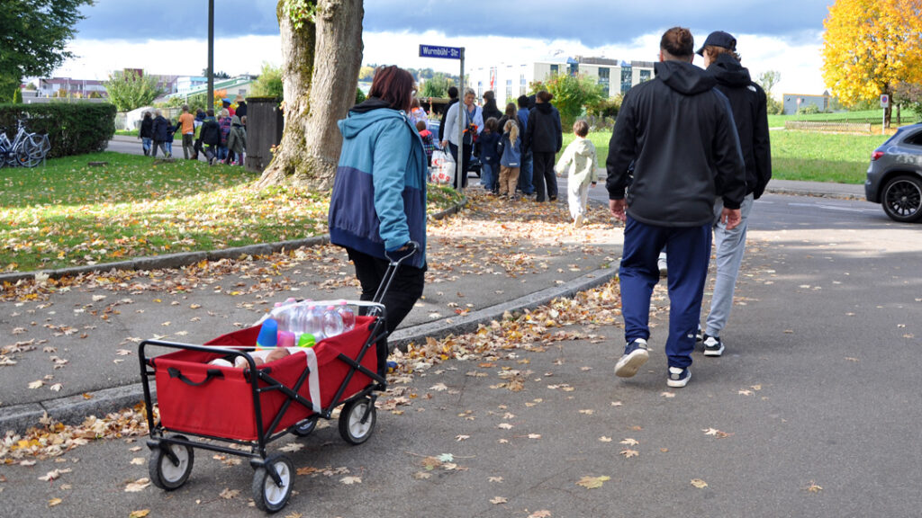 Leitende der Kinderspielwoche ziehen auf der Strasse einen Handwagen mit Brot, Bechern und Getränken. Vor ihnen gehen viele Kinder.