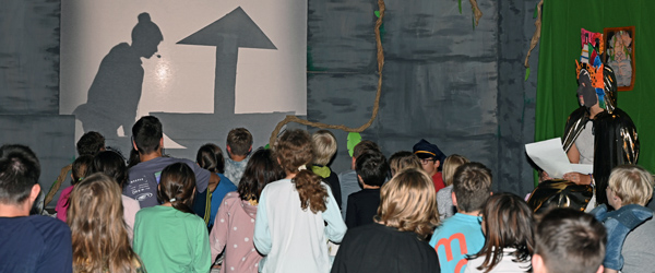 Kinder schauen ein Schattentheater mit einem Mädchen und einem Segelboot. Daneben sitzt ein Erzähler mit einer Maske im südamerikanischen Stil